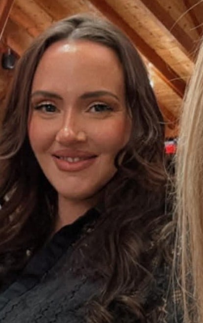 Portrait of a young woman with long dark wavy hair smiling at the camera, wearing a dark top with wooden background